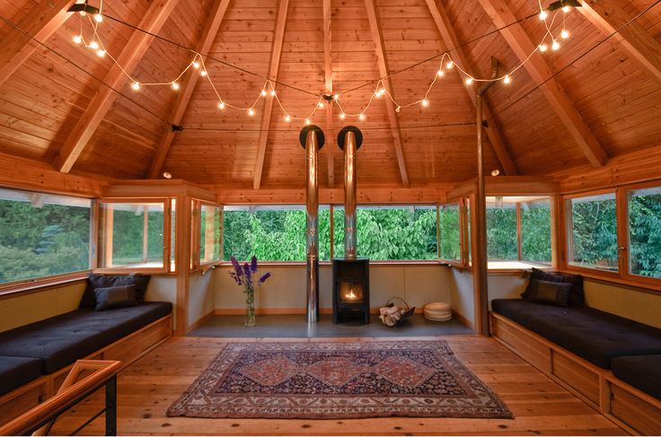 this is an interior shot of a woodpaneled cabin with windows, a rug, benches, lights, a stove, logs, flowers, pillows, cushions, and pipes. the cabin has a cozy atmosphere with a wood stove and windows looking out onto trees and nature. the furniture includes benches with cushions and pillows, and a rug in the center of the room. the lighting is provided by string lights hanging from the ceiling, which gives the room a warm and inviting ambiance.
