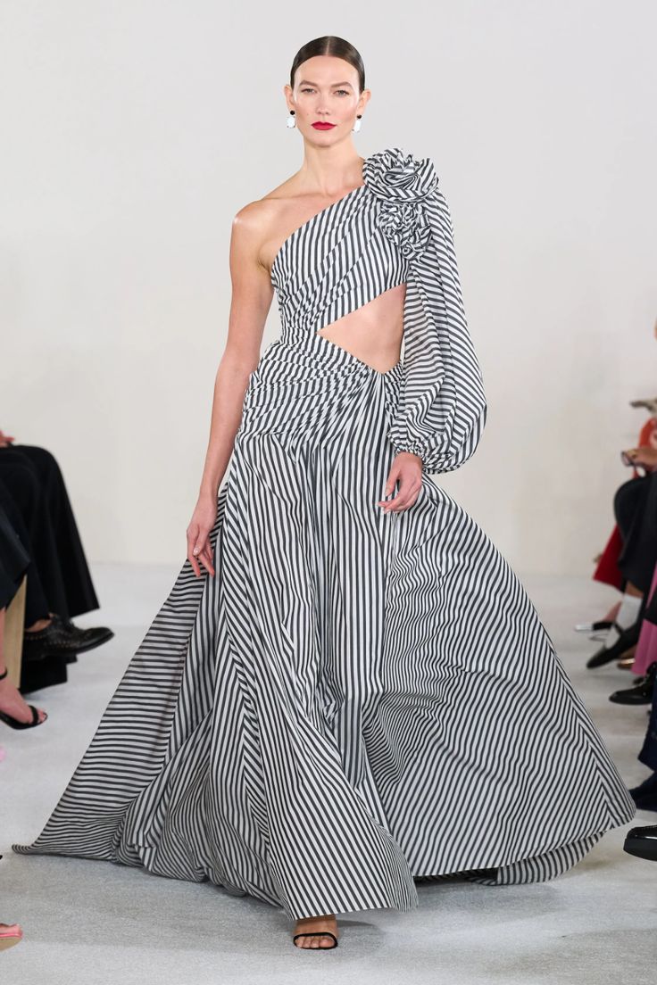 a female model walks the runway in a black and white striped one shoulder dress with a long sleeve. the dress has a cut out at the waist. the model is wearing black heels and has her hair pulled back. the background is blurry and there are other people in the background.