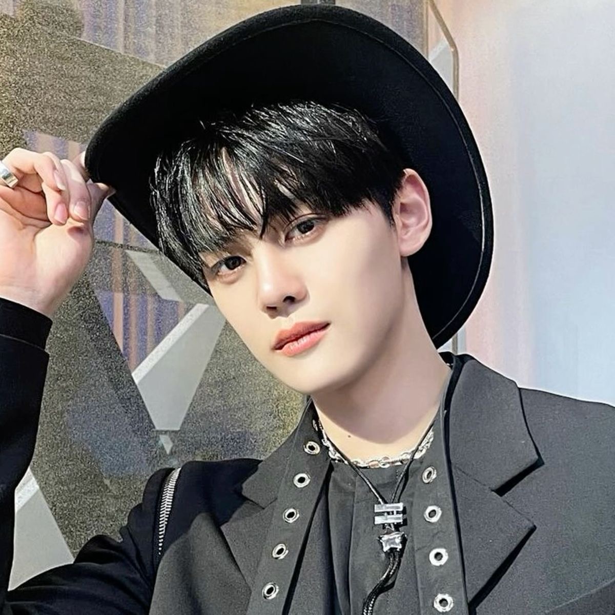 a closeup selfie of a young asian man wearing a black cowboy hat and a black suit jacket. he is accessorizing with silver rings and a silver necklace with a pendant. he has short black hair and is posing against a gray and white patterned wall. the image appears to be taken in a studio setting with professional lighting.