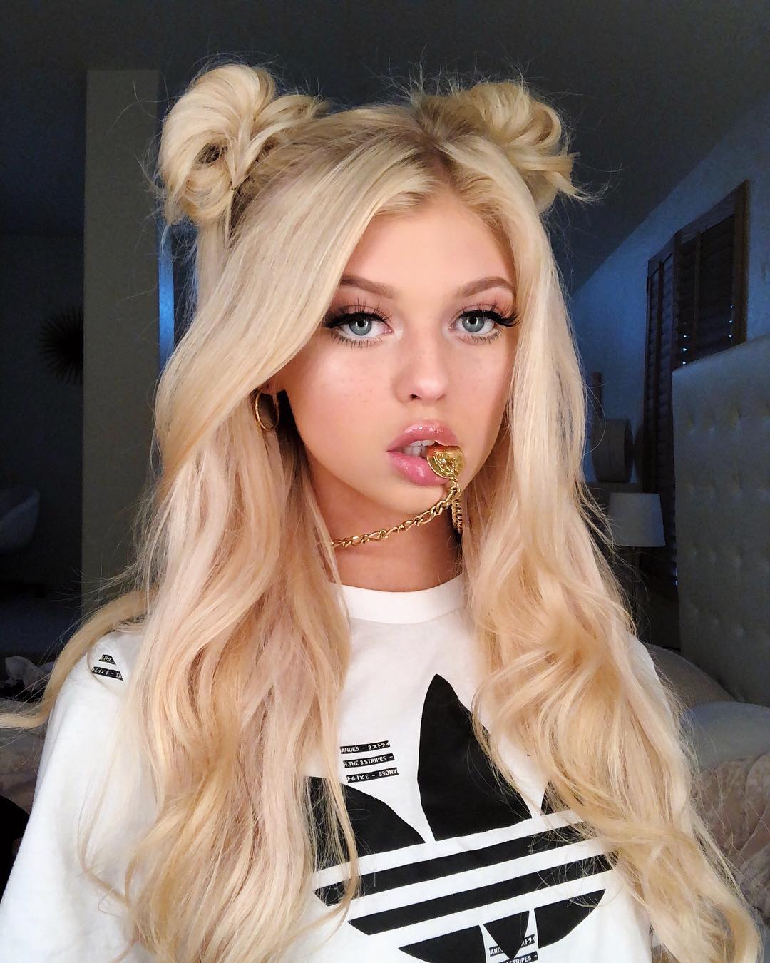 a portrait of loren gray with blonde hair styled in space buns, wearing a white adidas tshirt and a gold chain in her mouth, with blue eyes and glossy lips.