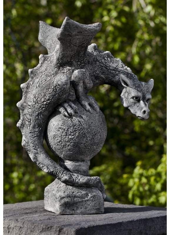 a stone statue of a dragon perched atop a ballshaped ornament, with a leafy green background. the dragon has textured skin, wings, and claws, and a slightly menacing expression. the statue is weathered and gray, giving it an aged appearance. it sits on a base that appears to be part of a larger stone structure.