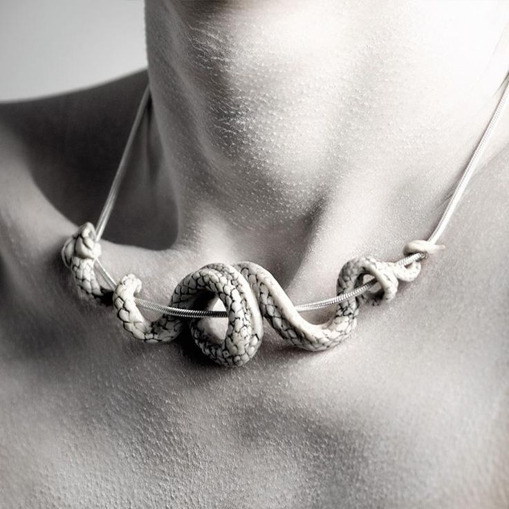 a monochrome closeup shot of a person's neck and upper chest wearing a snakeshaped necklace. the necklace features a detailed, coiled snake design, suspended from a thin cord. the snake's scales are visible, adding texture and detail to the jewelry piece. the image is focused on the necklace and the person's skin, creating a striking and artistic composition.