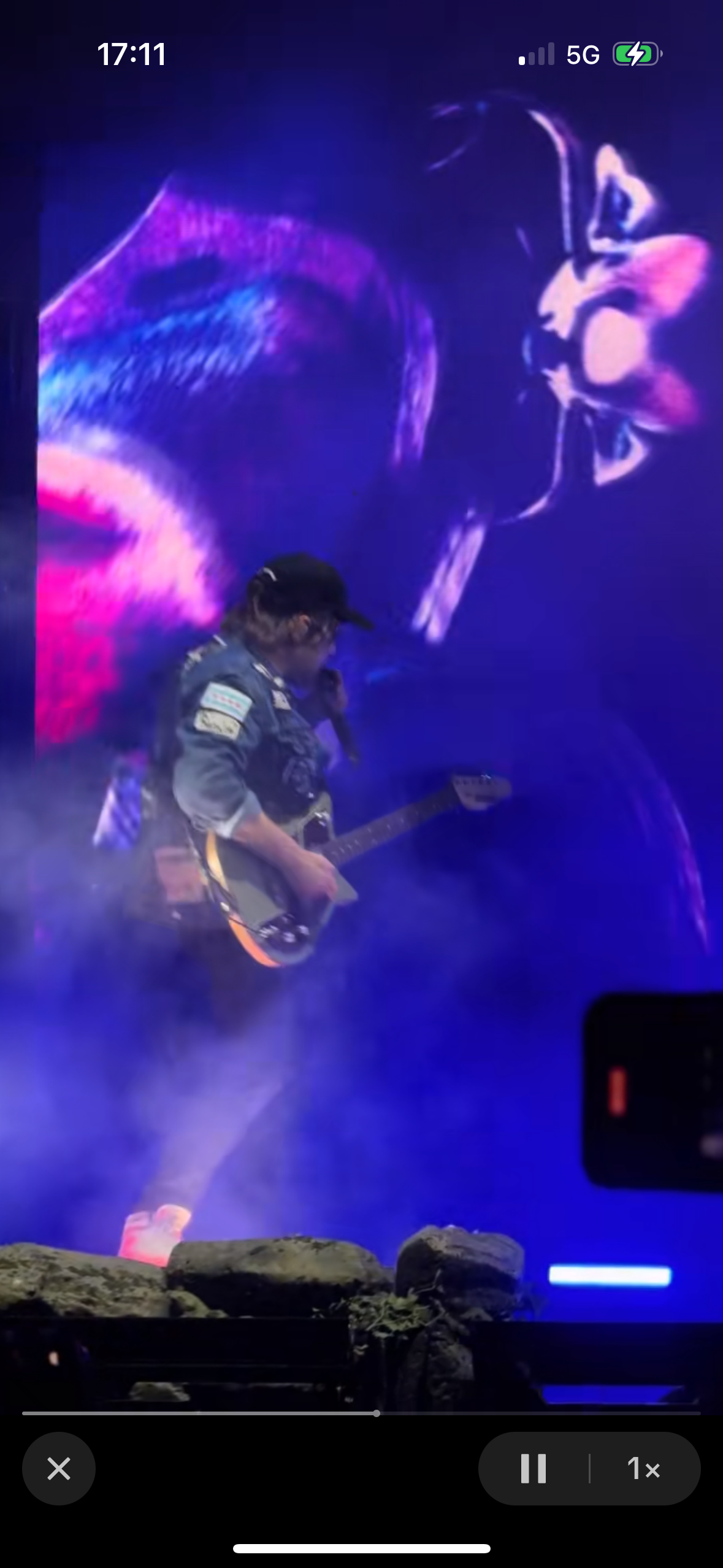a man wearing a black cap and a blue jacket is standing on stage playing a guitar. he is in the middle of a live performance with bright lights shining on him. the stage is dark and there is a faint image of a person behind him.
