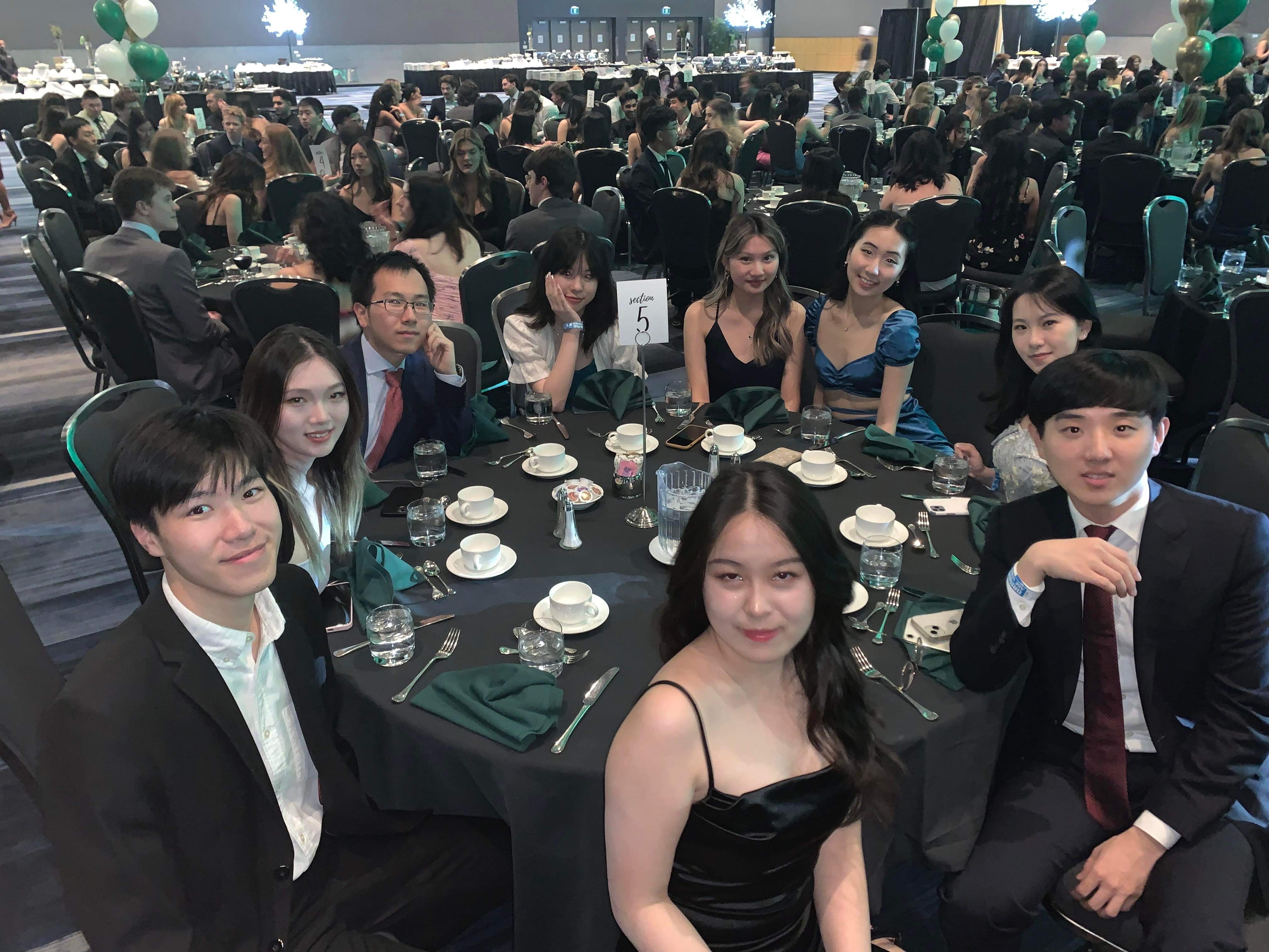 group of ten young adults are sitting at a round table covered in black cloth, smiling for a photo. they are dressed in formal attire, suggesting they are attending a celebratory event like prom or a graduation banquet. the room is large and decorated for the occasion, with round tables filling the space. the photo is taken from a slightly low angle, and the lighting is artificial.