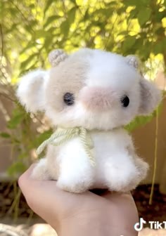 a small, white plush toy that looks like a baby cow is being held in a hand. the toy is very soft and fluffy with brown spots on its face. the toy has big, brown eyes and a green scarf around its neck. the hand is holding the toy up against a background of green leaves.
