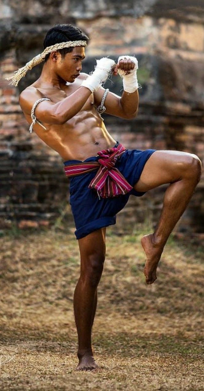 a muscular man, likely a muay thai fighter, is captured in a dynamic pose.  he's wearing traditional muay thai attire: a mongkol (headband),  shorts, and hand wraps. his body is lean and toned, showcasing the physical demands of the sport. the background features the weathered ruins of an ancient structure, suggesting a historical or cultural context. the image is sharply focused, with vibrant colors and strong lighting, emphasizing the power and skill of the athlete. the overall impression is one of strength, tradition, and the artistry of muay thai.