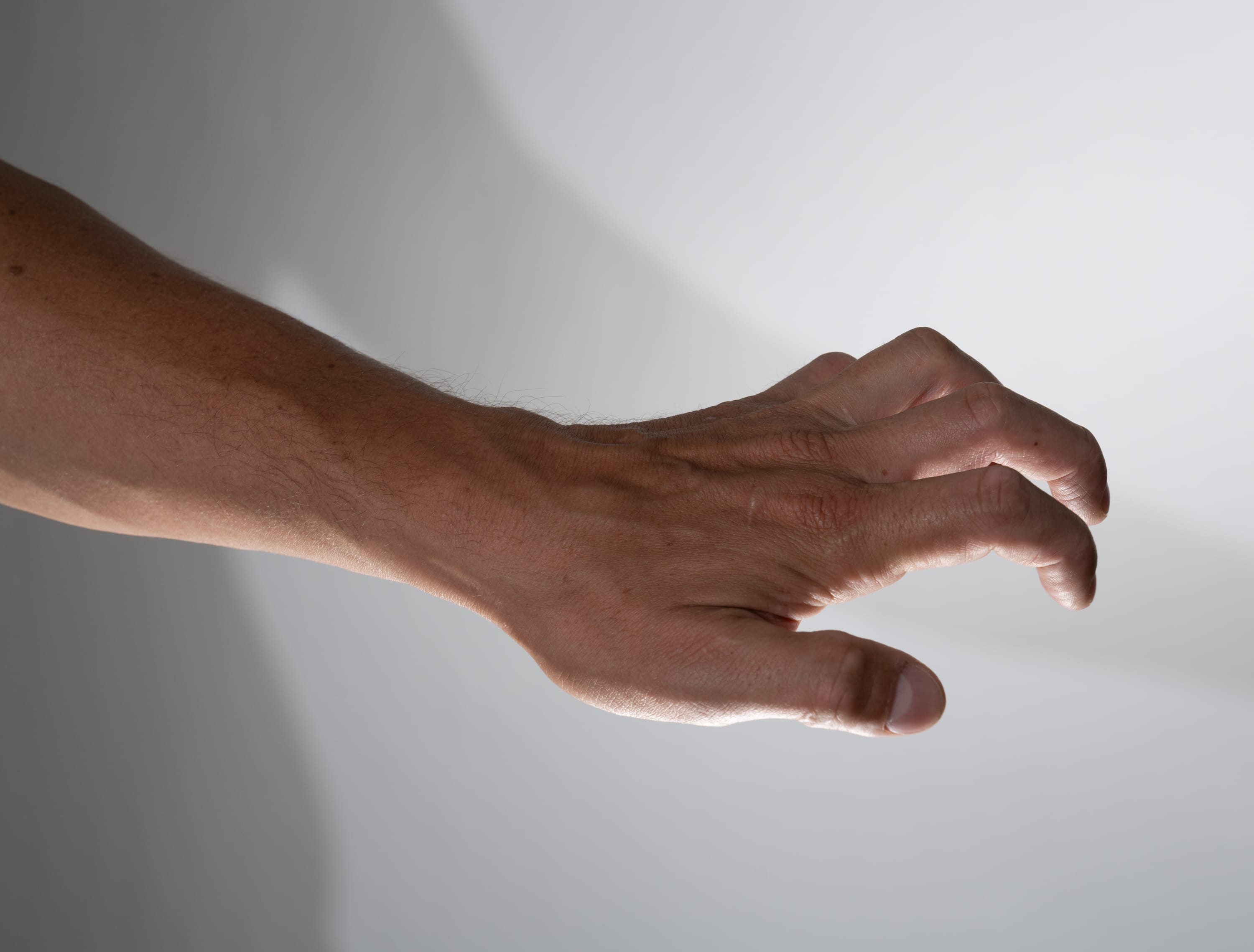 closeup side view of a man's outstretched arm and hand, with fingers slightly curled, against a light gray background with subtle shadows. the hand appears to be reaching or grabbing towards something out of frame. the lighting is soft and natural, highlighting the texture and veins of the skin.