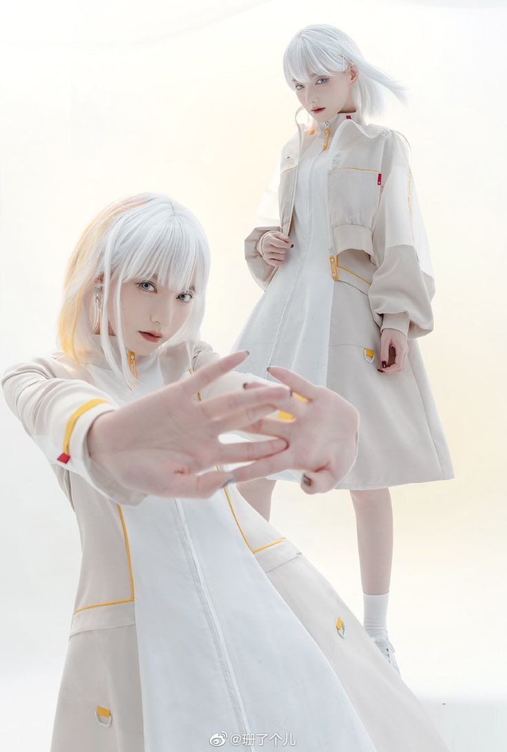 two women with white hair are wearing white dresses with yellow accents and posing for a photoshoot in front of a white background.