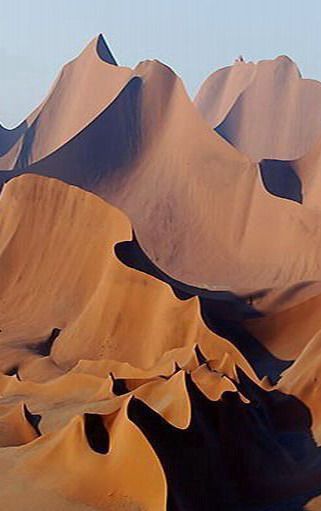 a wide shot of a huge orange sand dune. the desert is a rich orange and the sand is textured with ridges and dips. the desert appears to be endless.