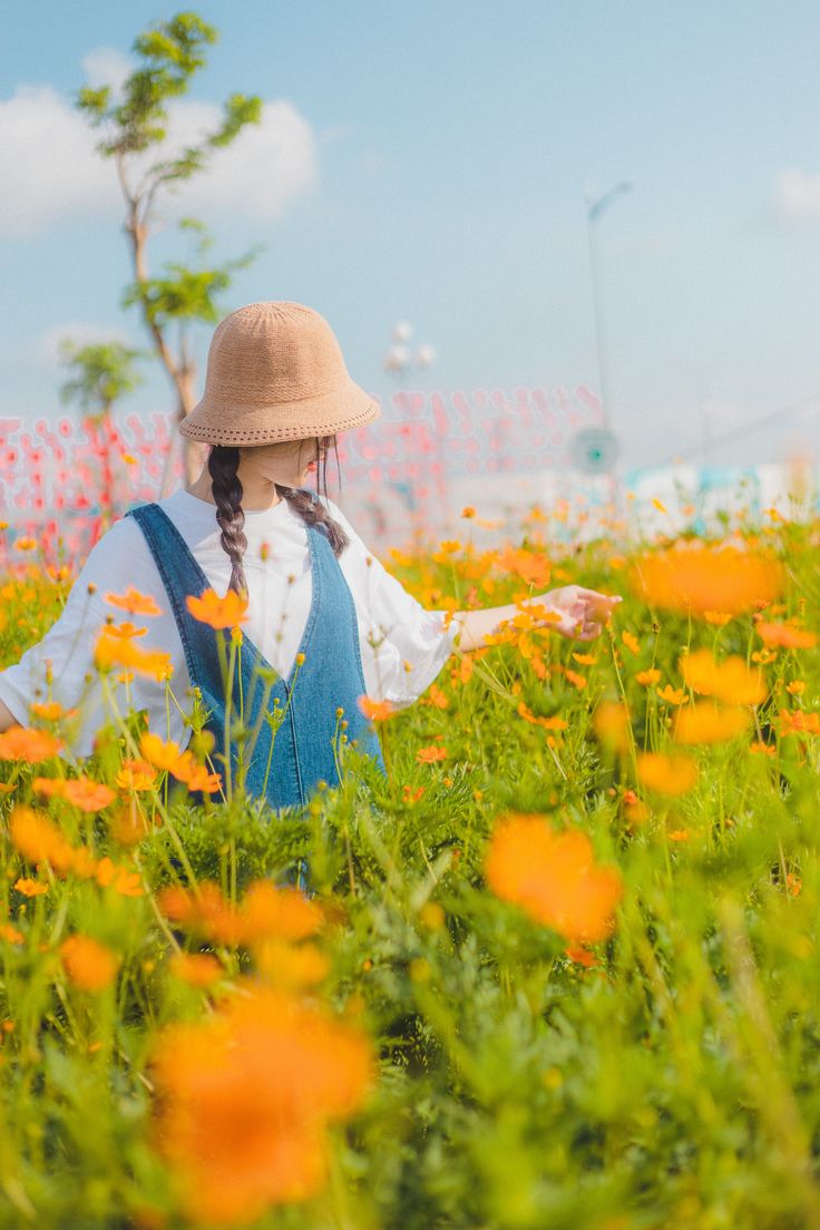 a young woman with pigtails wearing a straw hat and denim overalls stands in a field of vibrant orange cosmos flowers. the scene is bright and sunny, with a soft focus on the flowers in the foreground and the woman in the midground. the overall mood is peaceful and serene.  the woman appears happy and carefree.  the image is a portrait style shot and evokes a sense of idyllic beauty.
