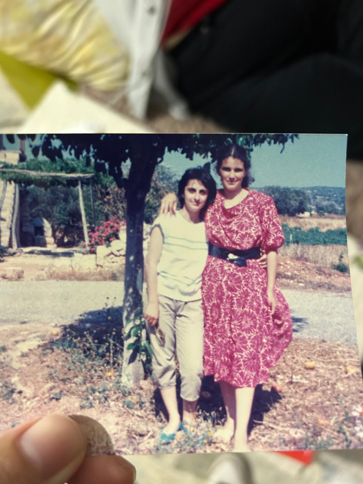 a closeup shot of a hand holding a vintage photograph. the photo depicts two young women standing outdoors in what appears to be a mediterranean setting.  one woman is wearing a pink floral dress, and the other is wearing beige pants and a lightcolored shirt. they are standing under a tree, and the background shows a rural landscape. the photo appears to be from the 1980s, based on the fashion and style. the women seem happy and close, suggesting a moment of friendship.  the overall tone evokes nostalgia and memories of the past.