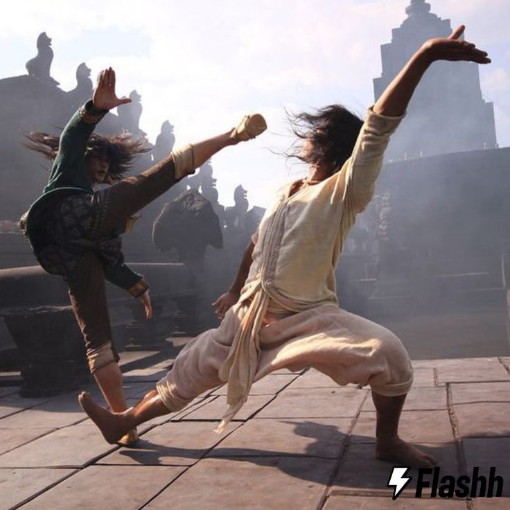 two female martial artists are captured in midfight against a backdrop of ancient temple ruins. the woman on the left is kicking with her right leg, while the woman on the right is blocking with her arms and leaning back. the image is a dynamic snapshot of their combat. the scene appears to be taking place on an outdoor set for a film or tv show. the overall sense of the image is actionpacked and exciting.