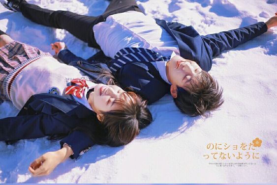 a young couple in school uniforms are lying in the snow, smiling brightly.  the boy is wearing a blue blazer and grey pants, and the girl is wearing a navy blue blazer and plaid skirt. the background is white snow, and the sun is shining brightly. the text in the image is in japanese and translates to "this dandelion flower, please give it to you".