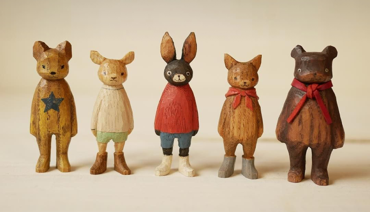five small wooden animal figures stand in a row against a cream background. from left to right: a yellow bear with a star, a light brown deer with green shorts, a black rabbit with a red shirt, a light brown fox with a red scarf, and a dark brown bear with a red scarf. the figures have a simple, folk art style and are painted in muted colors.