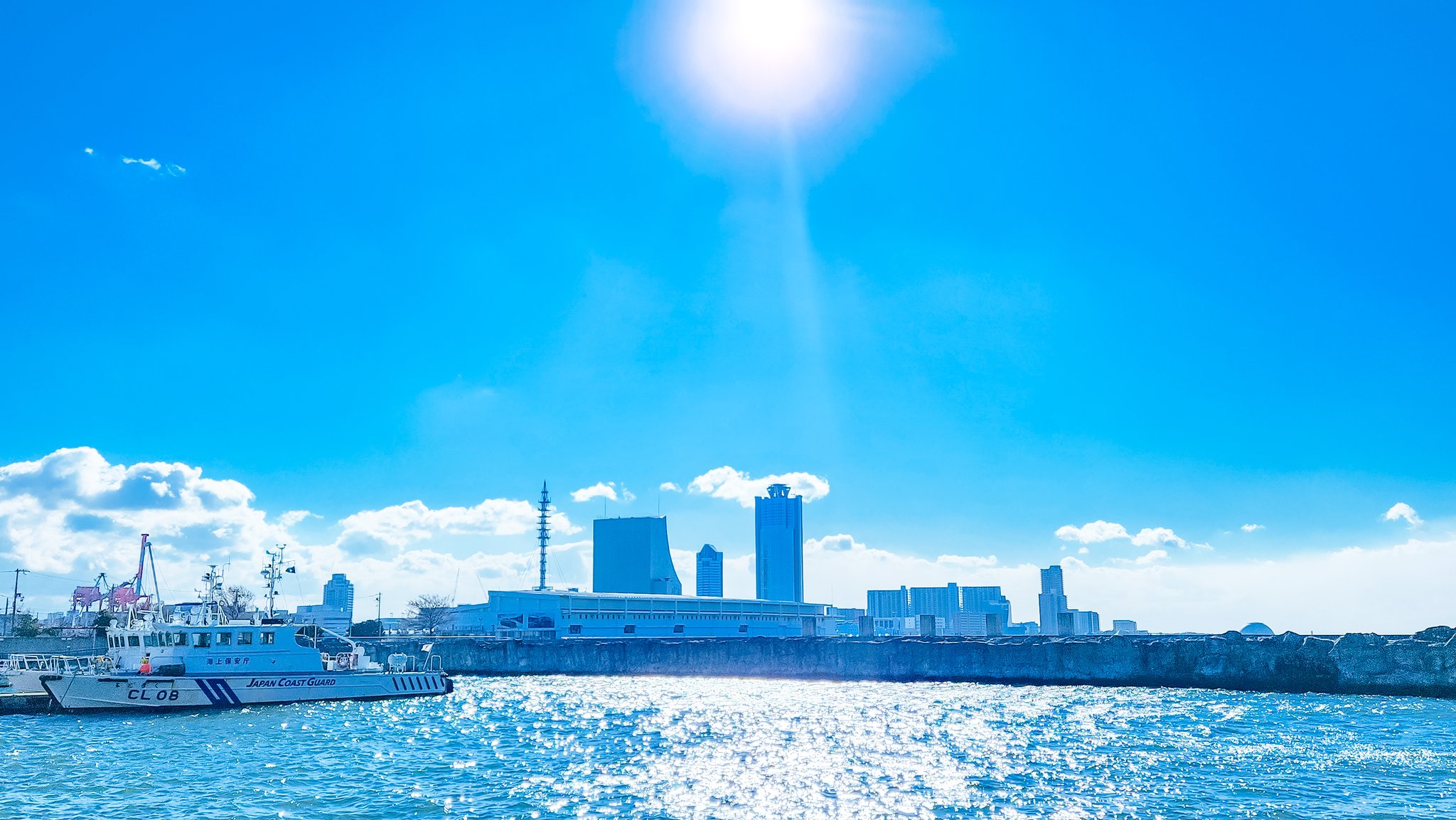 a wide shot of a sunny day, with a bright blue sky and a low horizon line. there are white clouds dotted around the sky. the foreground is taken up with water, with the sun's reflection shimmering in the centre. two ships, a coast guard boat and another, are to the left. a sea wall lines the coast, upon which sits a modern city of highrise buildings. to the left, an amusement park ride peeks above the buildings.