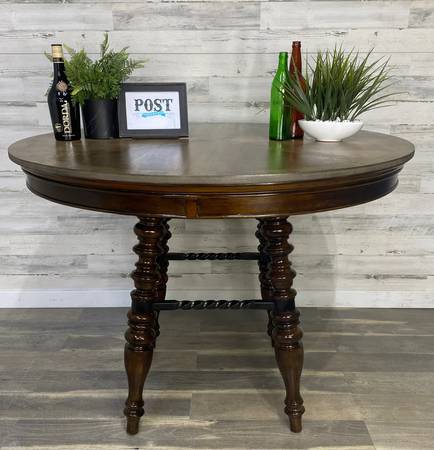 a round, dark wood table with ornate legs sits in front of a gray wash background and gray wood floor. on top of the table are several items, including a wine bottle, a potted plant, a picture frame with the word "post" written on it, and two more wine bottles with another potted plant.