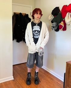 a young man stands in his room wearing a white cardigan, black shorts, and black boots. his hair is red and he has on several accessories.