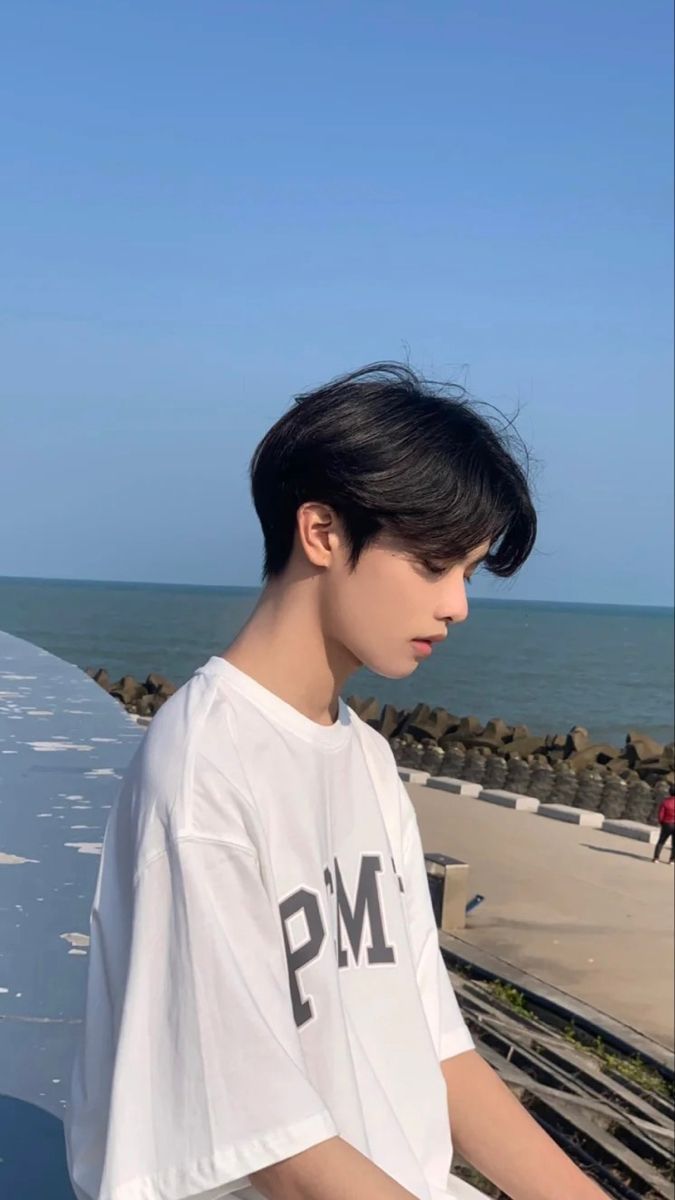 the image shows a young asian man standing by a seaside promenade. he is wearing a white oversized tshirt with the letters "pm" printed on it. the man has short, black hair styled with a middle part. the background features a clear blue sky, calm blue sea, and a concrete promenade. the image is taken from an eyelevel perspective and bathed in soft, natural light, creating a bright and airy atmosphere.