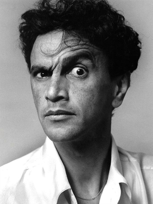 a black and white portrait of a man looking at the camera, with a serious expression on his face. he is wearing a white shirt with the collar up. the image is a closeup and the background is blurred.