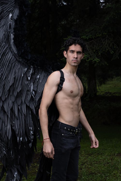 a shirtless man with a muscular physique stands in a dark forest, his back to a large pair of black angel wings. he's wearing dark jeans and has an intense gaze. the overall mood is dark, mysterious, and dramatic.