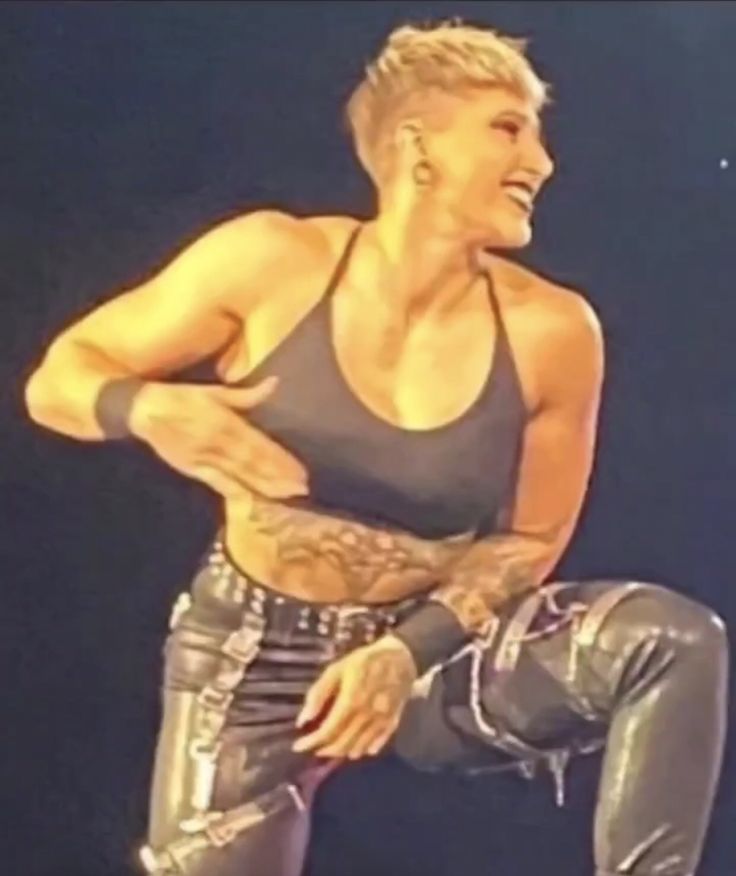 a photo of wrestler rhea ripley in wrestling gear with a black tank top, leather pants, and a short haircut, smiling on a wrestling stage. she is posing in the ring and appears muscular with tattoos visible on her arms and torso.