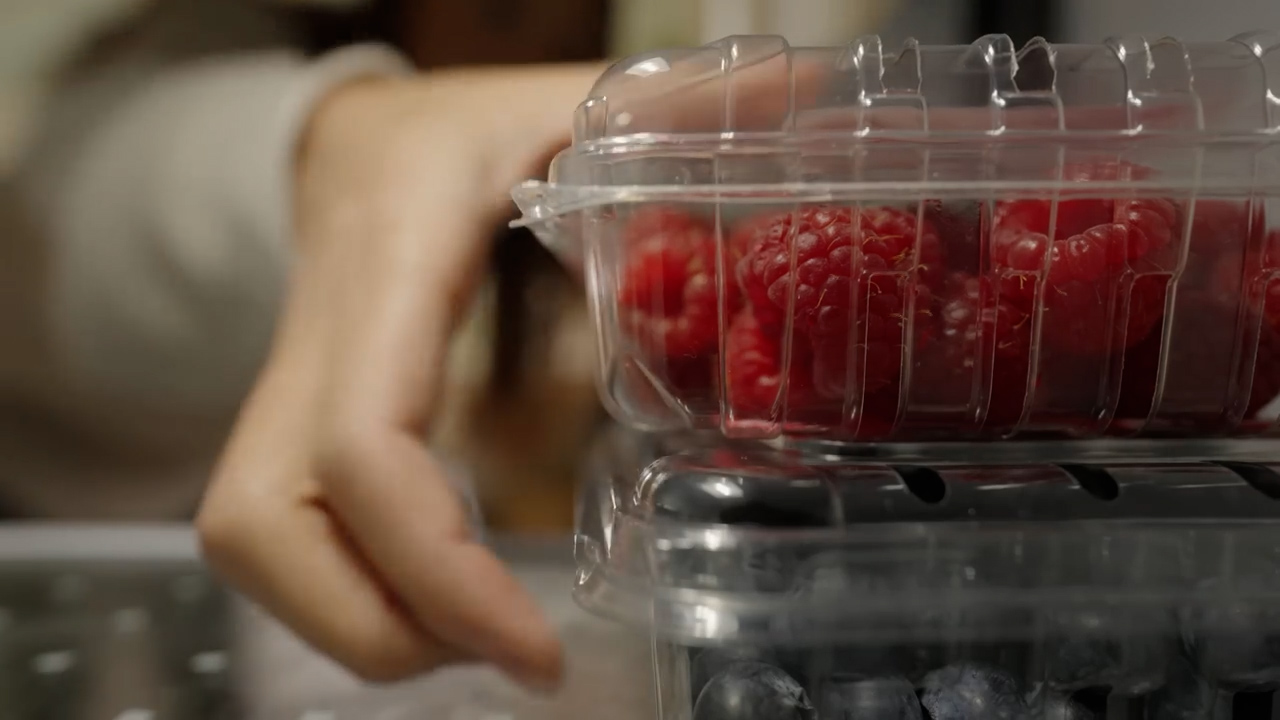 a closeup image of a woman's hand reaching for a clear plastic container of fresh raspberries. the container is stacked on top of another container filled with blueberries, both residing in a refrigerator. the focus is on the raspberries and the hand, creating a sense of anticipation and desire for a healthy snack.