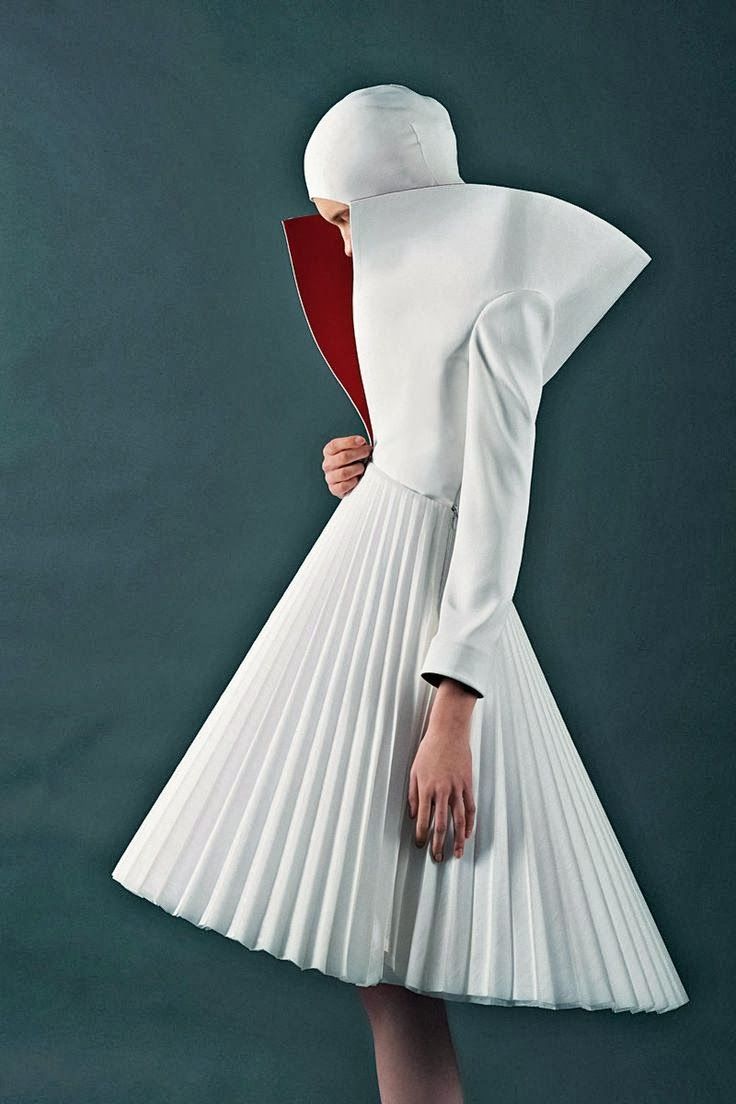 a woman in a white pleated dress with a red collar and a hood stands in a studio, facing away from the camera. her face is partially obscured by the hood, and her hands are clasped together in front of her. the dress is long and flowing, and the pleats are very defined. the background is a simple blue wall. the overall effect is of a minimalist and elegant fashion photograph.