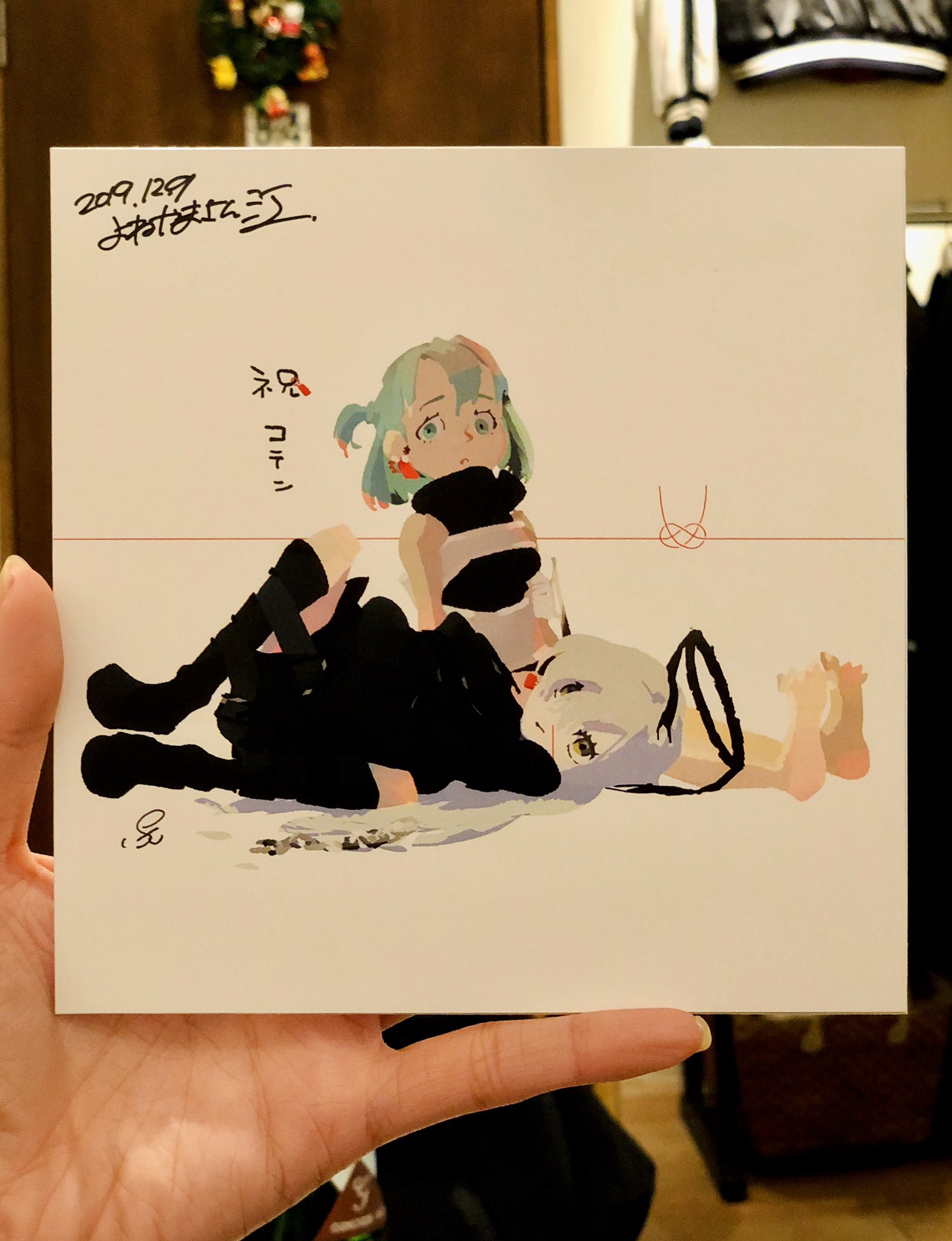 the image shows a drawing on a white paper held by a hand. the drawing features two animestyle female characters in casual clothing lying down on their sides, with one girl's head resting on the other's back.  there is japanese text and a signature on the top left corner of the paper.