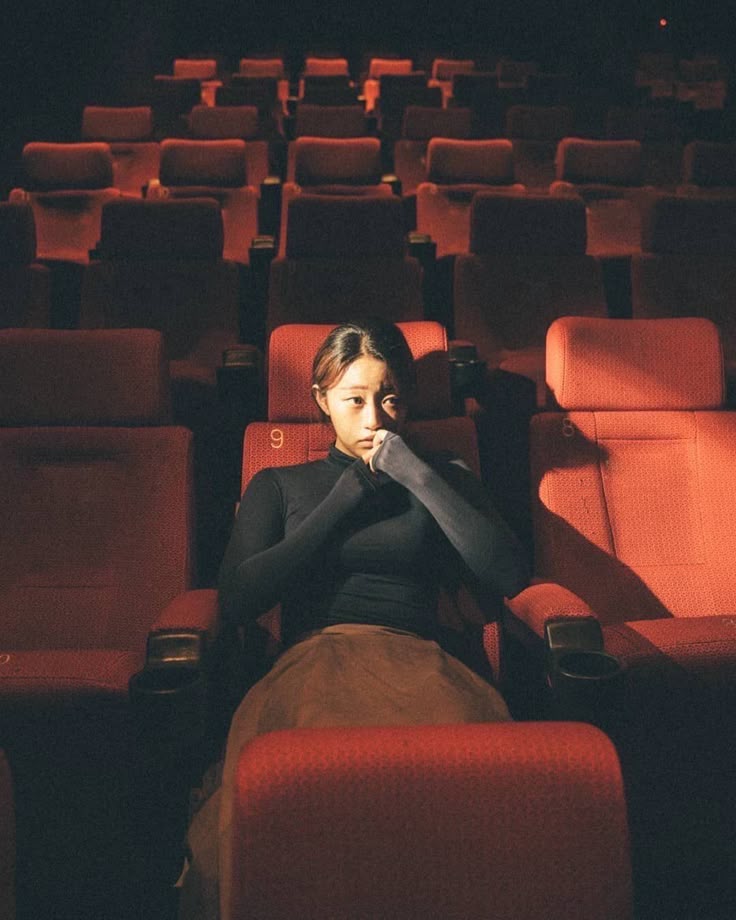 a young woman sits alone in a mostly empty movie theater. she's wearing dark clothing and has her hand to her face, looking pensive. the seats are red, and the background is dark.