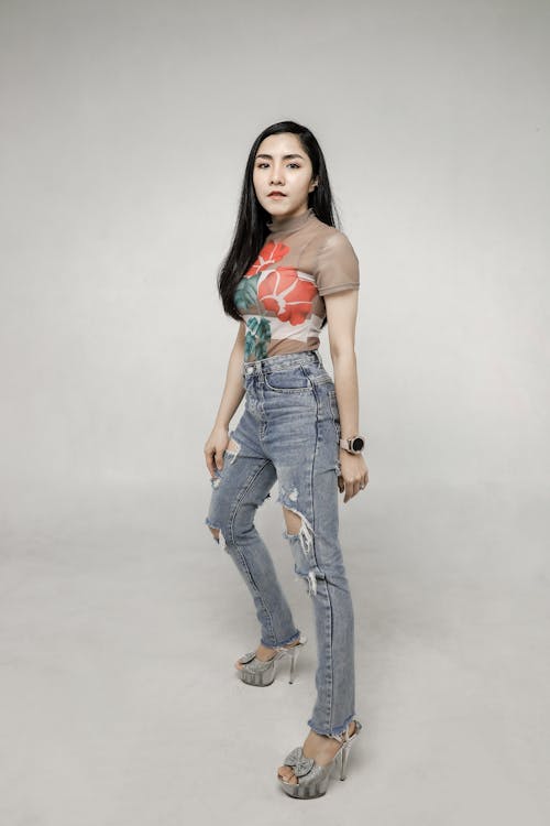 a full body studio shot of an asian woman. she is wearing a floral top, ripped jeans, and platform heels. she also has a watch on her left wrist. she is standing in a slight stride with her right foot forward. the background is a light gray.