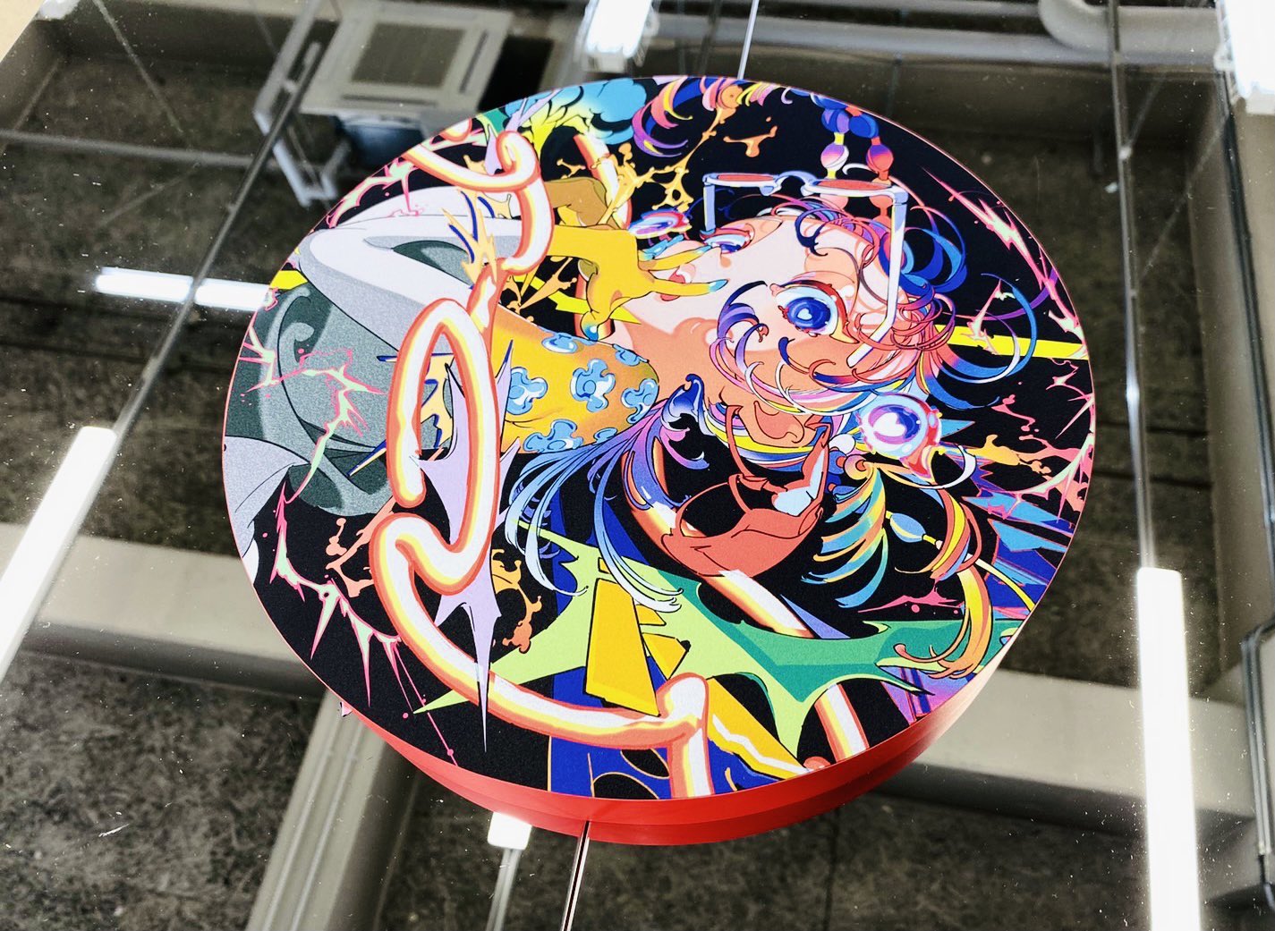 a round, brightly colored illustration of a girl with blue hair is hung from the ceiling. the artwork is framed by a red circle and is reflected in the ceiling above. the girl's eyes are wide and her expression is slightly manic. she appears to be surrounded by abstract shapes and lines in a variety of colors. the overall style of the illustration is modern and graphic.