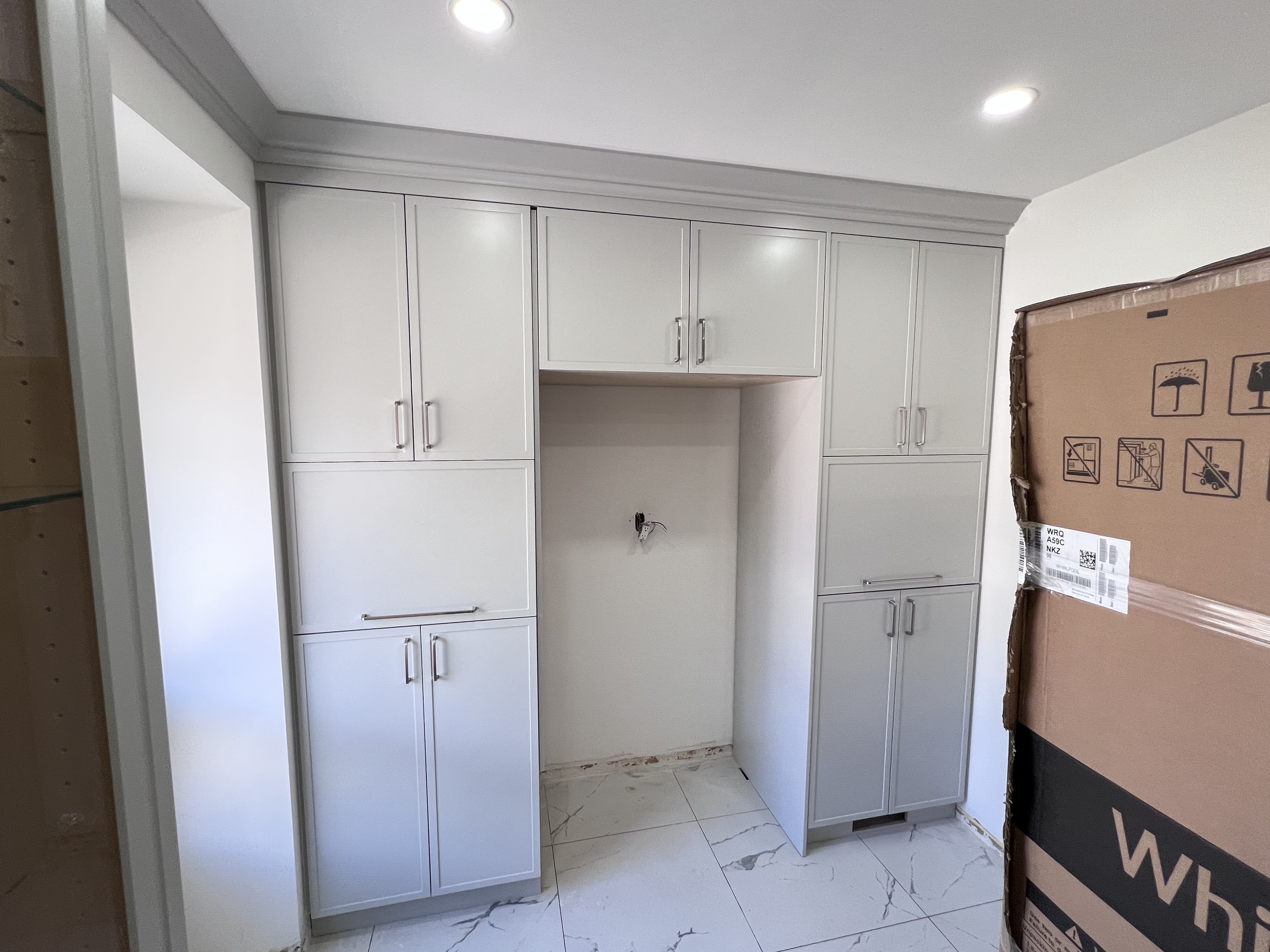 image shows a kitchen interior with newly installed grey cabinets flanking a space for a refrigerator. the cabinets are topped with crown molding. the floor is covered in white marble tiles. a large, unopened appliance box sits to the right. the overall scene suggests recent construction or renovation.