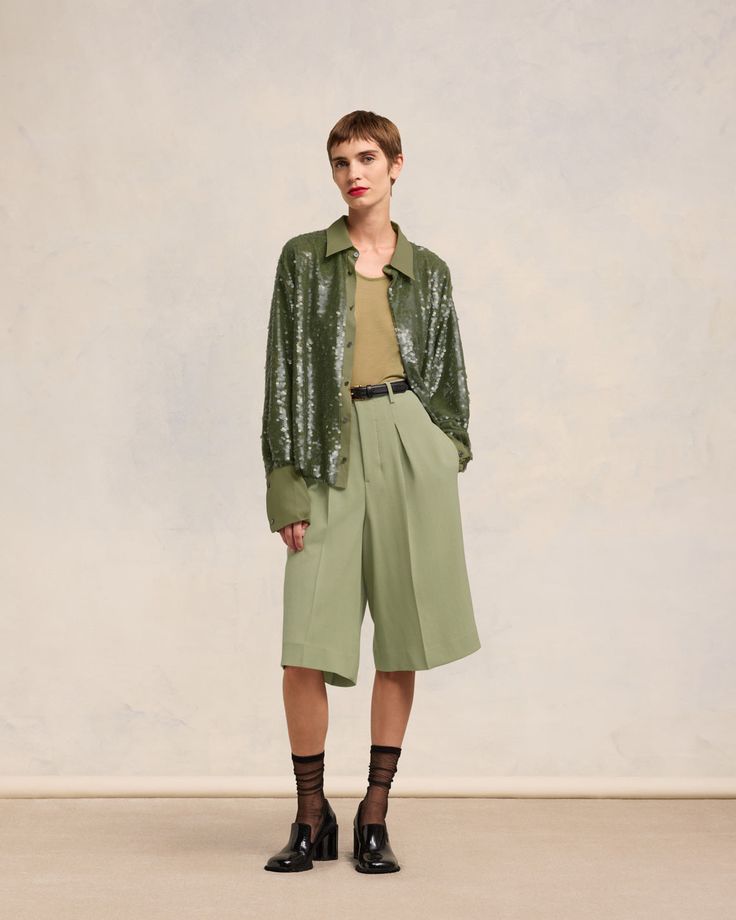 a full shot of a woman model standing against a neutral background. she is wearing a sage green sequined oversized shirt, light green highwaisted tailored bermuda shorts, black sheer socks, and black heeled shoes. her hair is short and styled. her expression is slightly serious. the overall style is elegant and modern, suggesting spring or summer fashion.