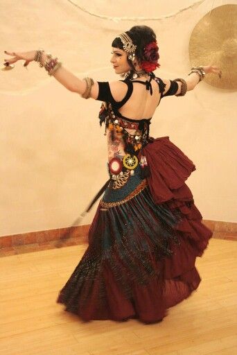 a belly dancer in a traditional costume is performing on stage. she has a red and blue skirt and many pieces of jewelry adorning her. she is wearing a lot of jewelry on her arms and head. her hair is dark and she is looking down at her hands as she poses.