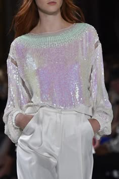 a woman in a white sequin top and white pants poses for a photo on a runway. she has red hair and her top is extremely sparkly. she looks straight ahead with a serious expression on her face. the background is blurry.