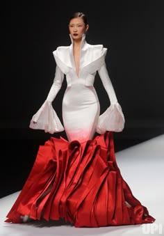 a woman in a white dress with red accents walks down a runway. the dress has a high collar and long sleeves. the skirt is a dramatic, red, ruffled design. the model is posing for the camera with a confident expression. the lighting is bright and the backdrop is simple, allowing the dress to be the focal point.