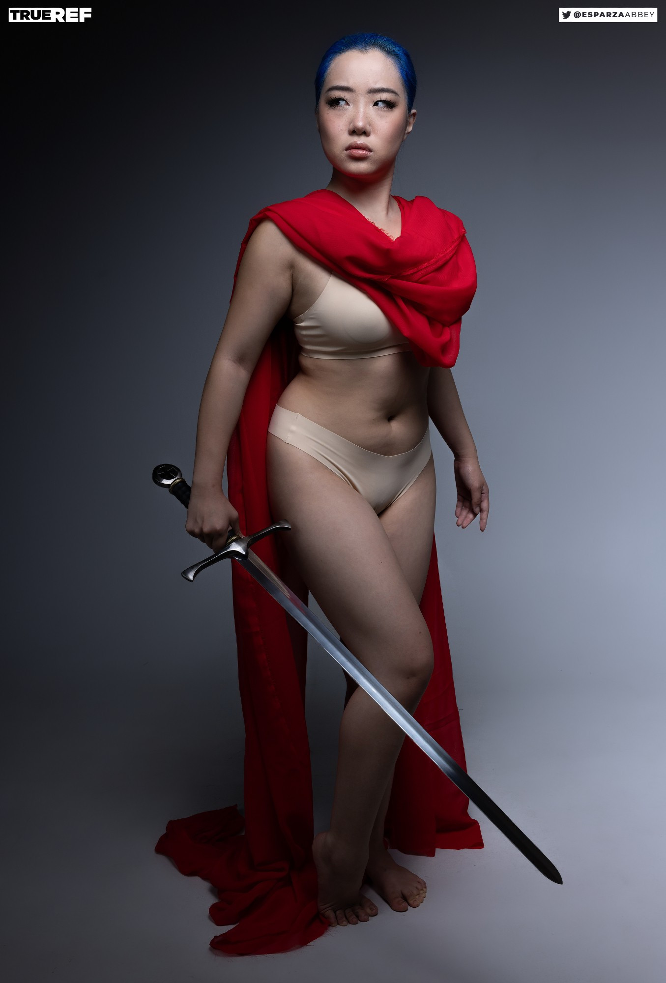 a fulllength studio shot of a woman with short, blue hair. she is wearing a red cape, beige underwear, and a beige bra. she is holding a sword and is posed in a strong and confident stance. the background is grey, and the lighting is dramatic. the overall mood is powerful and confident.