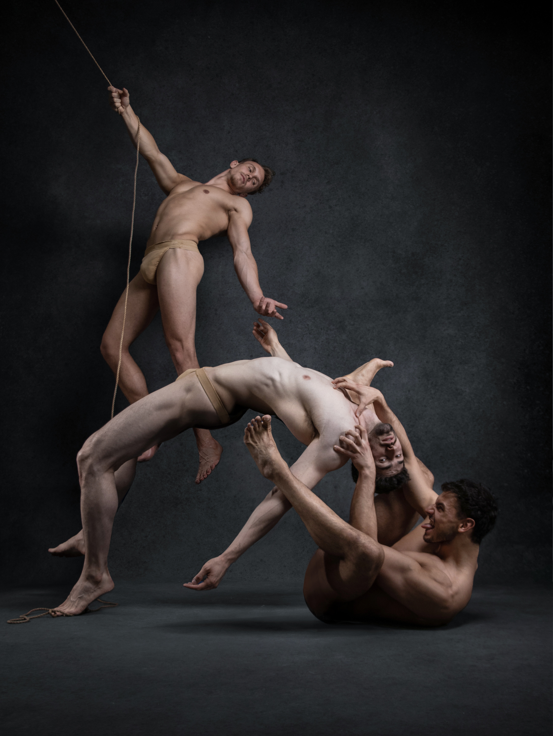 the image shows three male dancers in a dynamic pose against a dark background. one dancer is suspended in midair, held up by a rope. the other two dancers are intertwined on the floor below him, their bodies contorted in a display of strength and tension. the dancers are all nude except for fleshcolored briefs. the lighting is dramatic, highlighting the dancers' muscles and the lines of their bodies. the overall effect is one of power, grace, and athleticism.