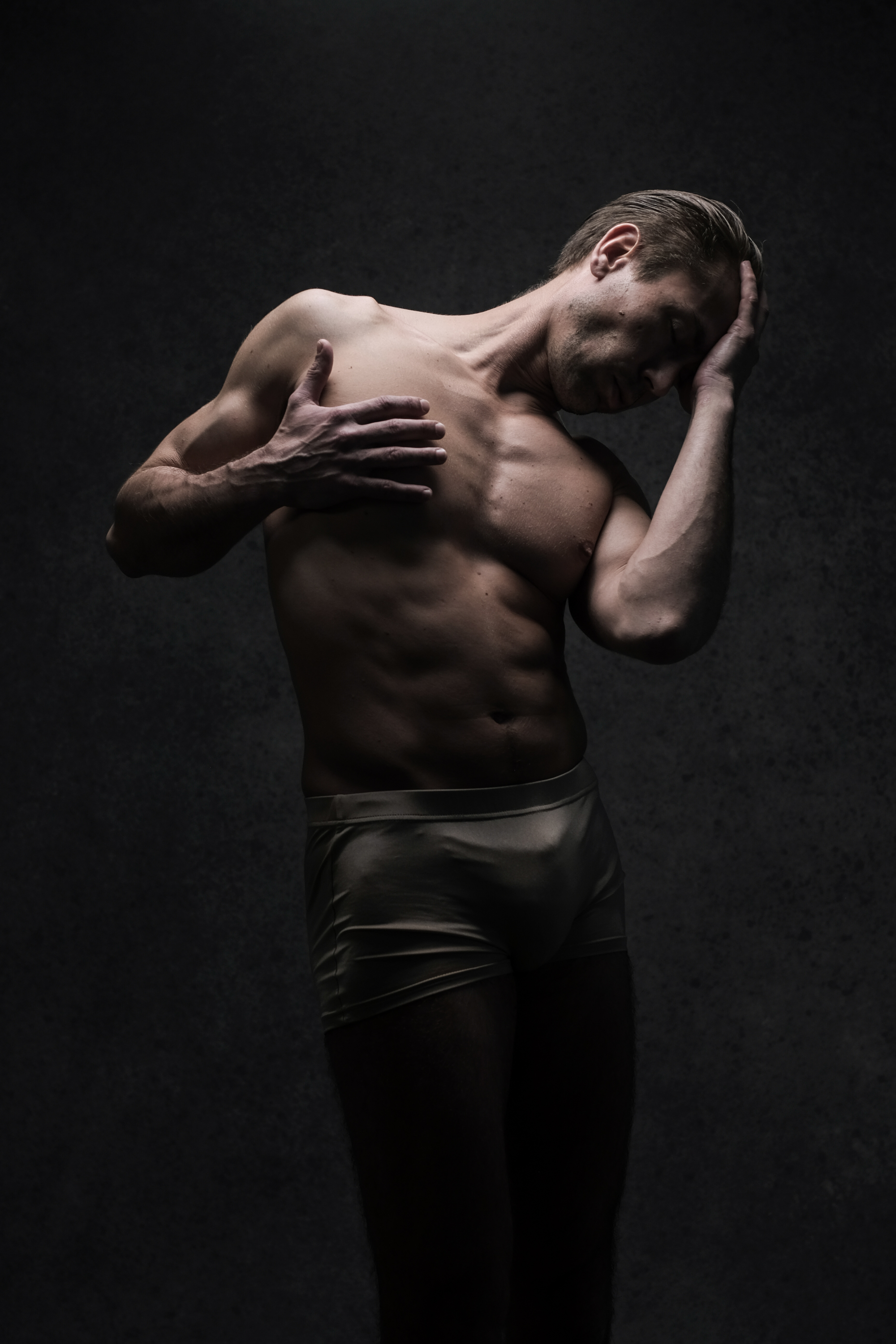 a man stands shirtless, wearing only underwear, against a dark gray background. he is lit dramatically, with deep shadows emphasizing his muscular physique. his head is tilted to the side and his hand rests on his forehead, creating a contemplative or melancholic mood.