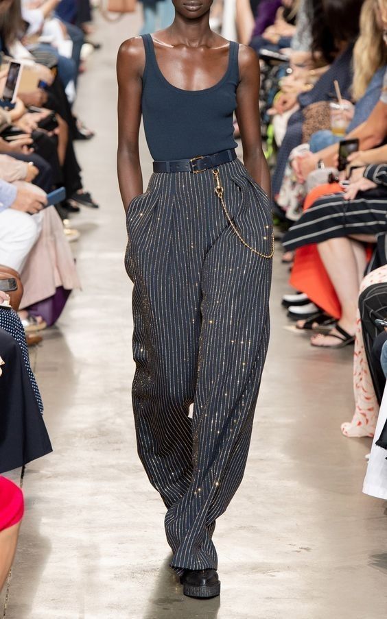 a female fashion model walks the runway wearing a black tank top, black and gold striped pants, and a black belt with a gold chain. the model is wearing black shoes with a white sole.  the runway is in a light gray color and there are people in the background.