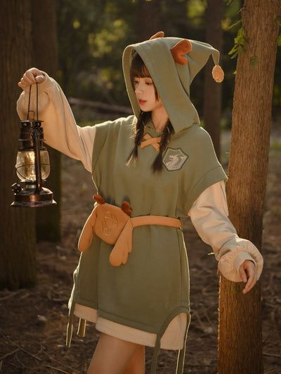 a woman stands in a forest holding an oldfashioned lantern. she is wearing a whimsical outfit that includes a green hoodie with attached reindeer antlers and pompom, a lighter colored tunic with puffy sleeves underneath, and a small fawnshaped pouch on a belt. the scene is lit by dappled sunlight filtering through the trees.