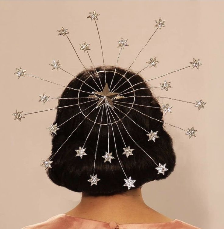 a woman with a bob haircut is facing away from the camera, wearing a silver headpiece with a large star in the center and smaller stars radiating outward on wires. she is wearing a pink dress. the background is a light beige color.