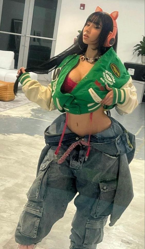 a woman with long black hair is wearing a green jacket, pink bra and blue denim cargo pants. she is posing for the camera.