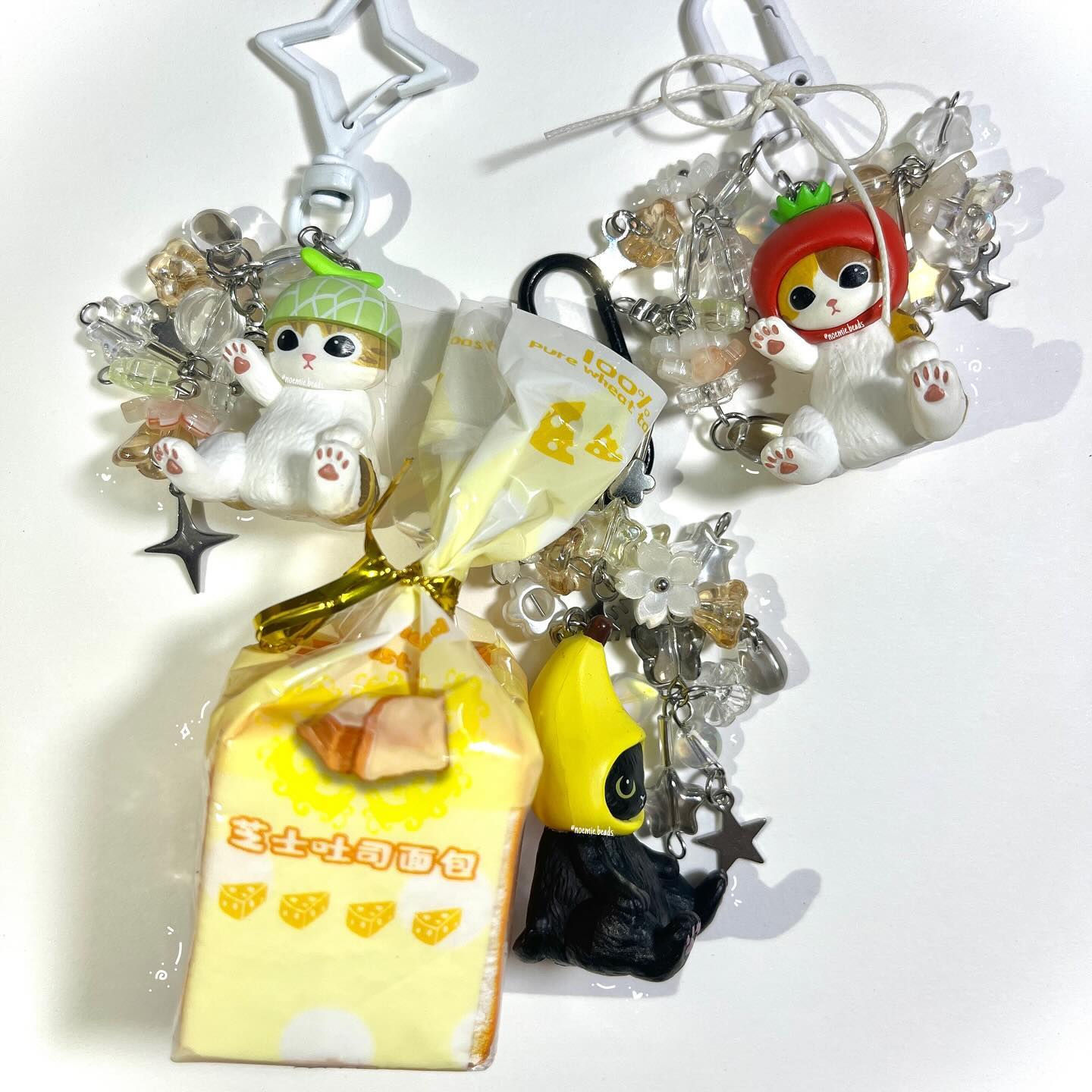 the image shows four keychains with cat charms. one cat is wearing an acorn hat, another a tomato hat, and a third a banana peel. there is also a keychain with a miniature loaf of bread with cheese decorations. all the keychains have beads and star charms.