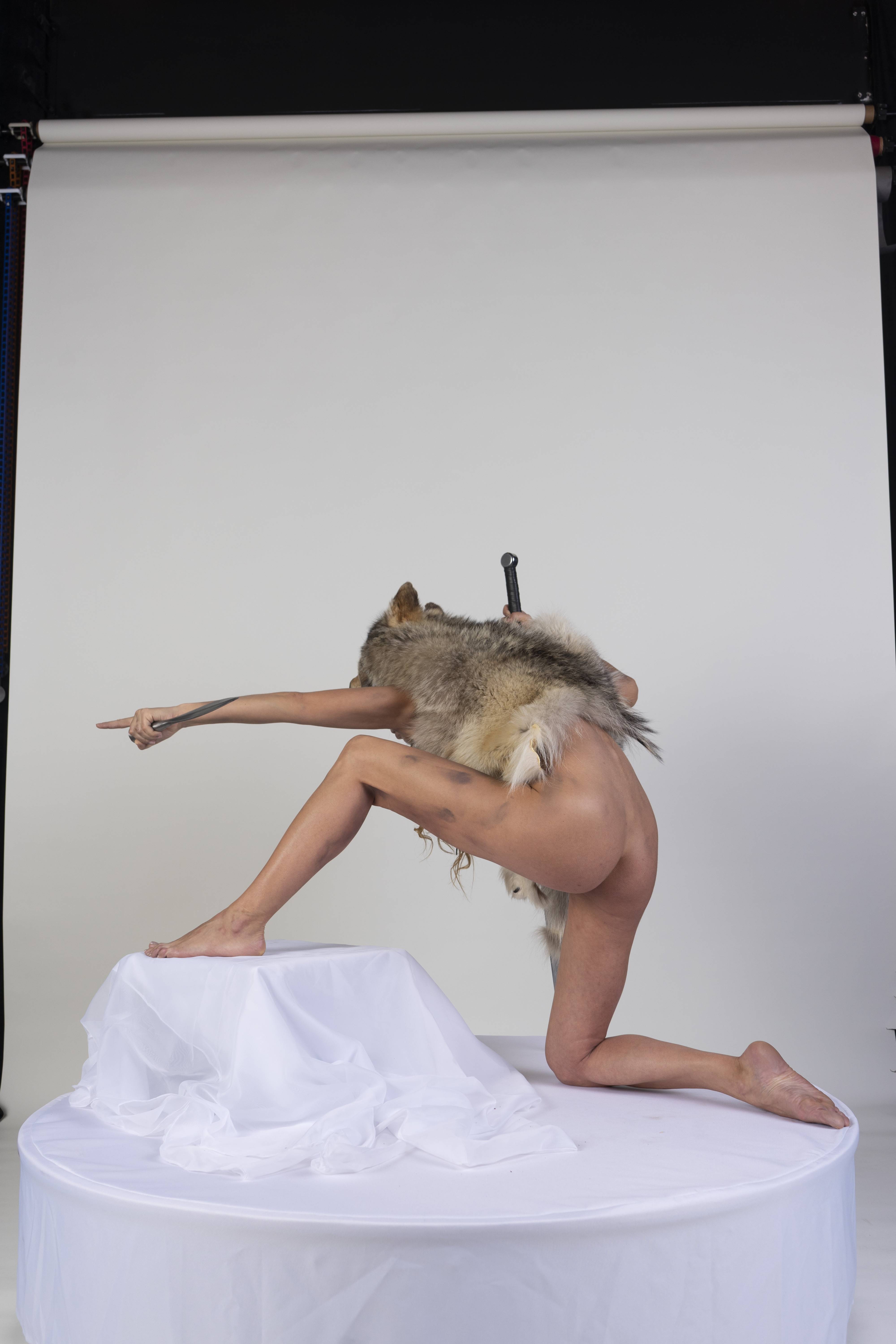 a nude woman is posed on a white platform with a white cloth draped over a small structure on top of the platform. she is wearing a wolf pelt over her shoulders and has a sword stuck in her back. she is pointing with her right hand. the background is a plain white studio backdrop.
