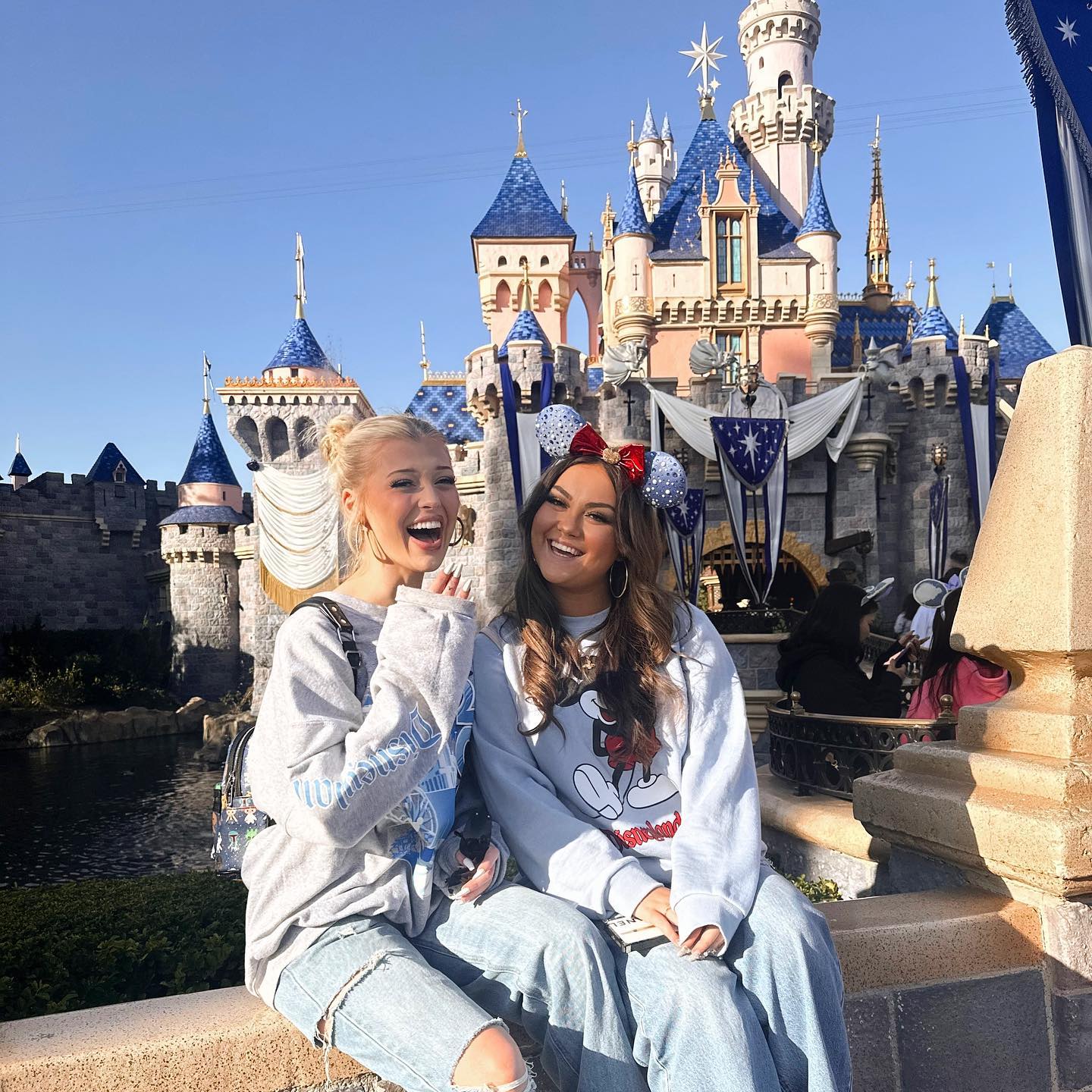 two women are posing for a photo in front of the disneyland castle. the castle is made of grey stone with blue and pink accents. both the women are wearing casual clothes; sweaters and jeans. the woman on the right is wearing minnie mouse ears with a red bow, and the woman on the left is laughing. the sky is blue, and the sun is shining.
