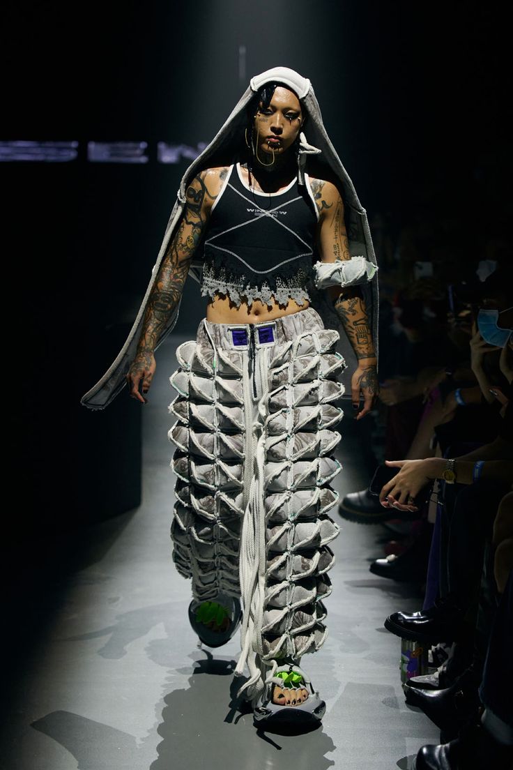 a fashion model walks down the runway wearing a unique and avantgarde outfit. the model is a woman with short black hair, wearing a grey hooded jacket, a black tank top with a white design, and grey pants with a distinctive design. she is accessorizing with a necklace and earrings, and her makeup is dramatic with dark eyeliner and a nude lip. the runway is dark and the audience is in the background. the image captures the bold and creative nature of fashion.