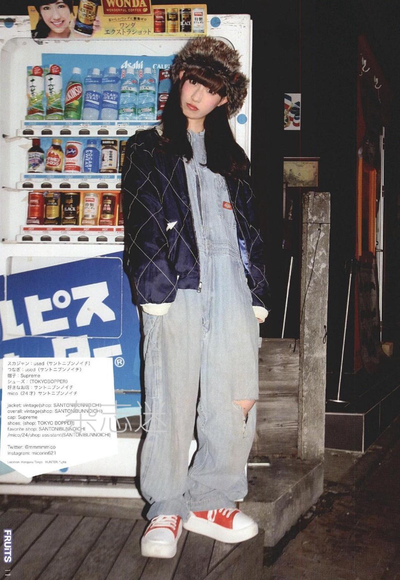 a young woman is posing in front of a brightly lit vending machine at night. she's wearing a black quilted jacket over light blue denim overalls, a black beanie hat, and red and white platform sneakers. the image has a vintage, filmlike quality with muted colors.