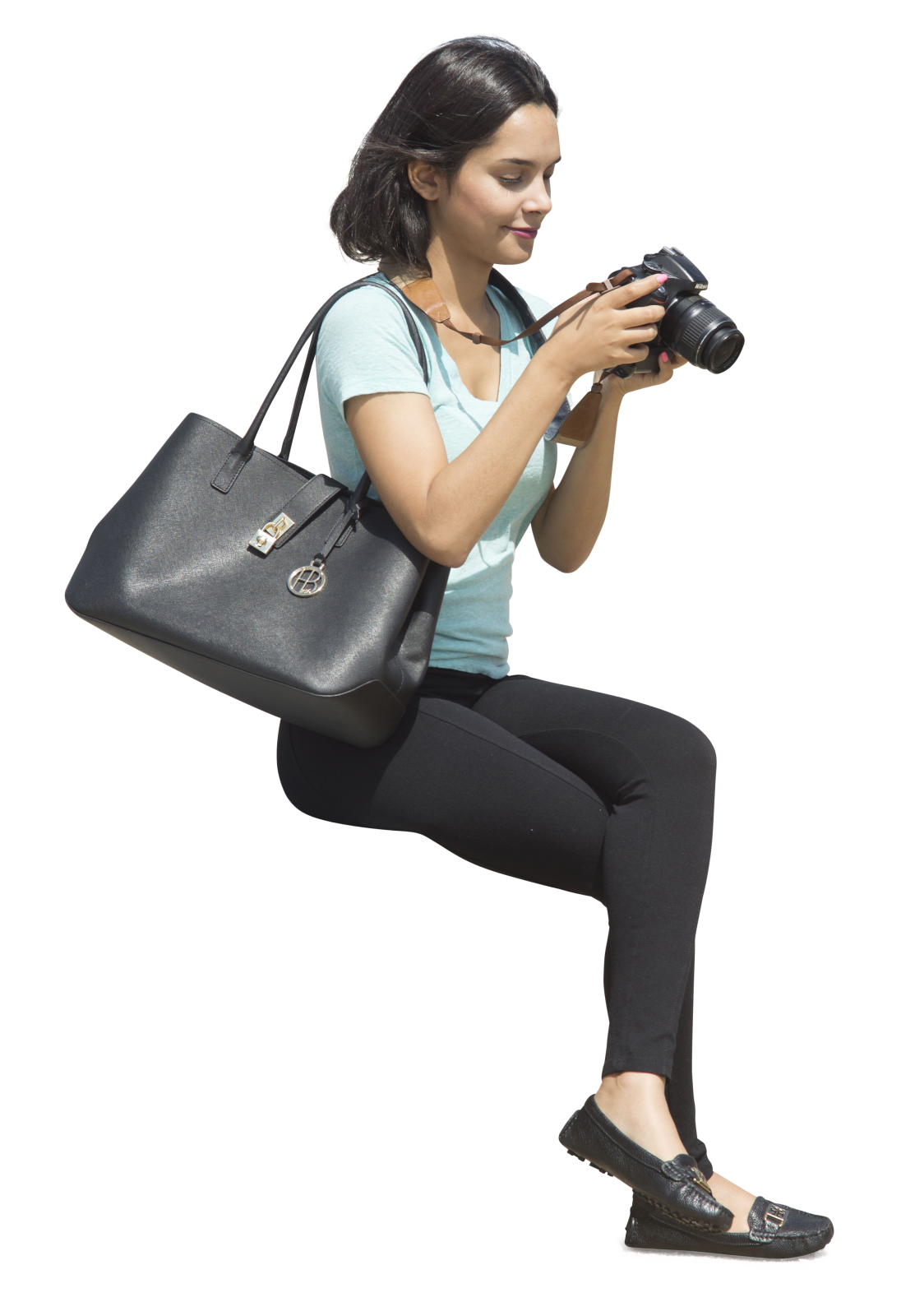 a woman with shoulderlength black hair is sitting and looking at a camera in her hands. she's wearing a light blue tshirt, black pants, and black flats. she's also carrying a large black tote bag. the background is removed.