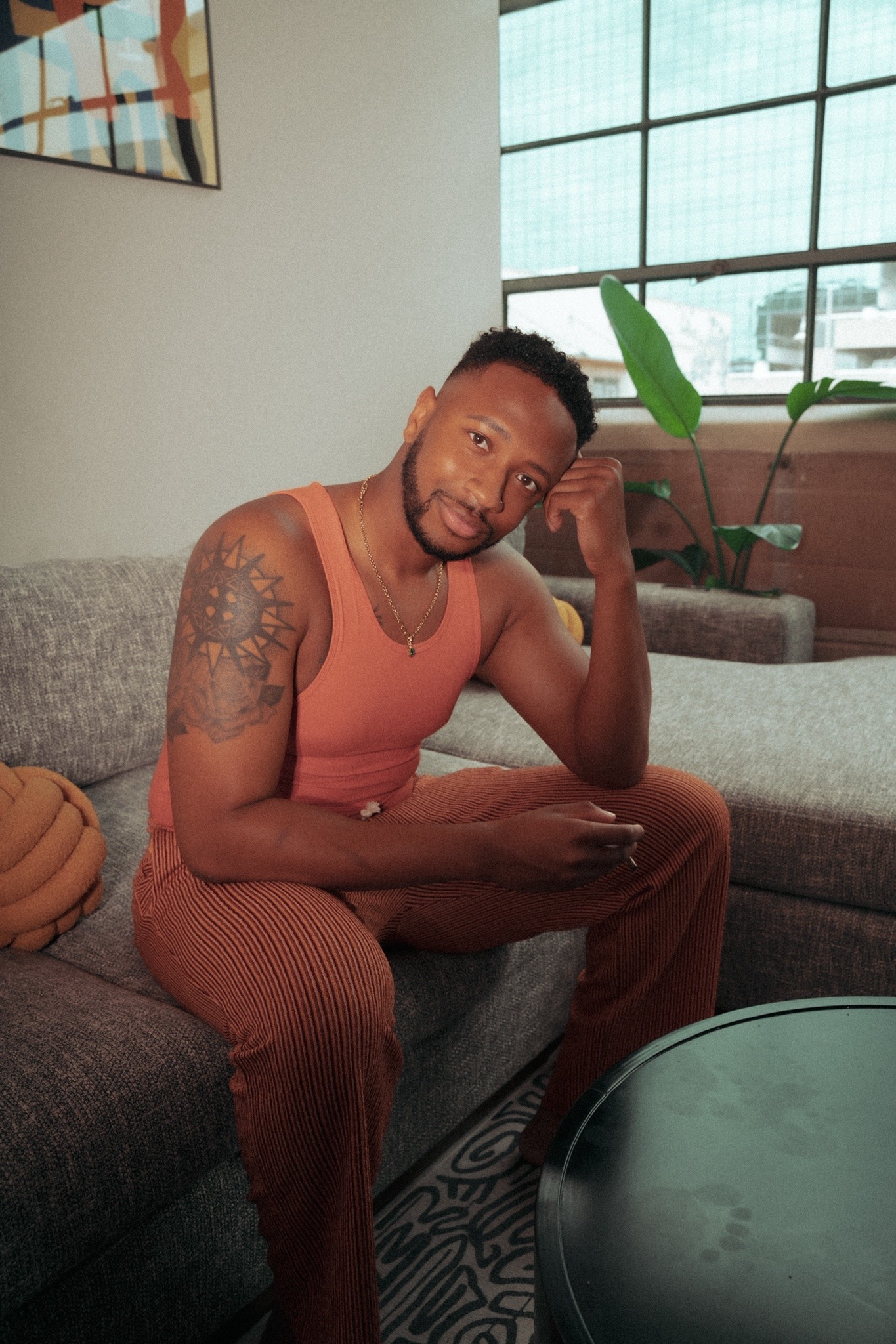 a man with brown skin and black hair sits on a grey couch and looks directly at the camera with a slight smile. he is wearing a light pink tank top and brown corduroy pants. his left hand is resting on his chin and his right hand is on his lap. there is a small rug under his feet. in the background, there is a plant in a pot on the other side of the couch, and there is a window behind him that is closed. there is a coffee table to the right of the man with the edge of it shown in the image.